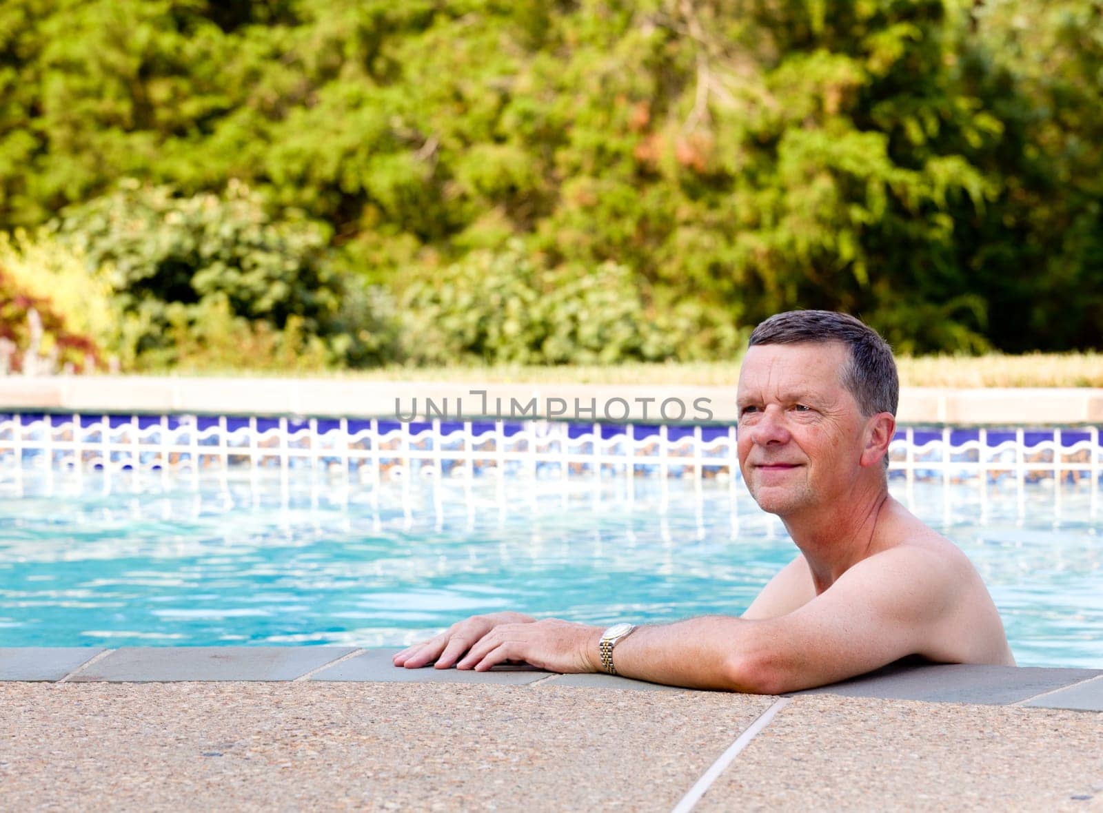 Senior male relaxing by the side of a modern swimming pool in back yard garden and facing the camera with smile