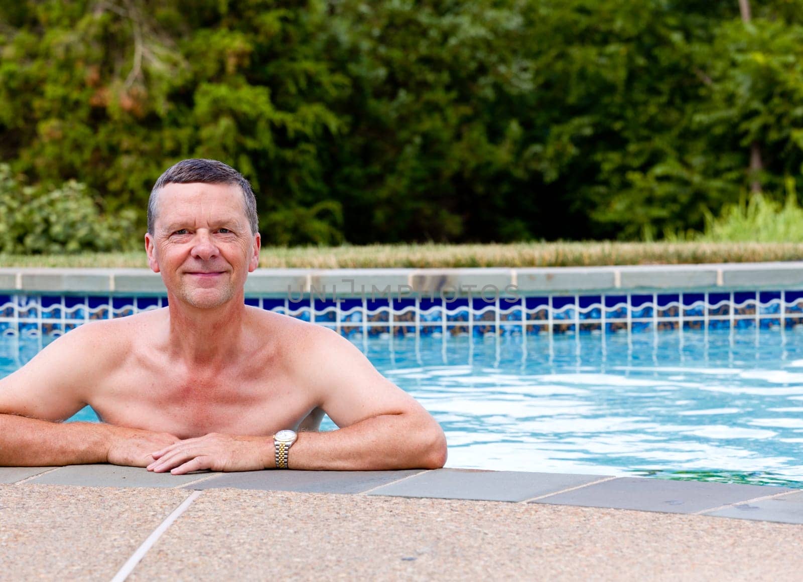 Senior male relaxing by the side of a modern swimming pool in back yard garden and facing the camera with smile