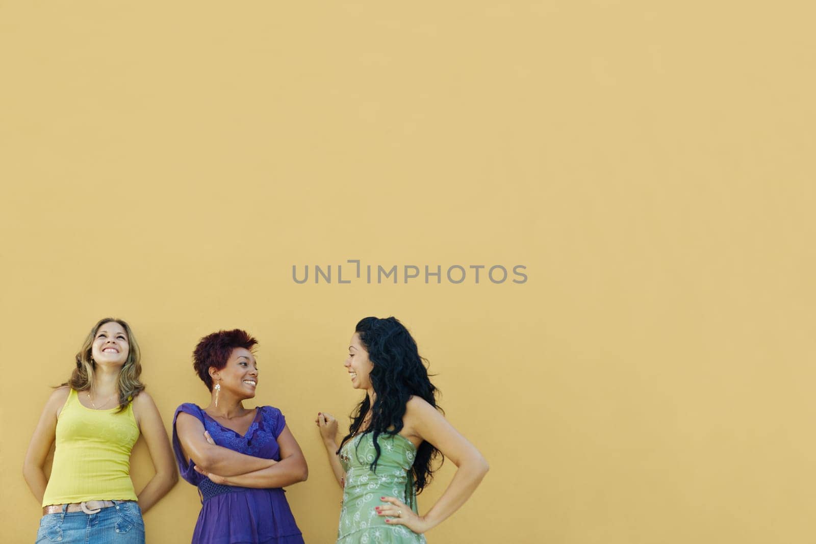 group of three female hispanic friends leaning on yellow wall and laughing. Horizontal shape, waist up, copy space