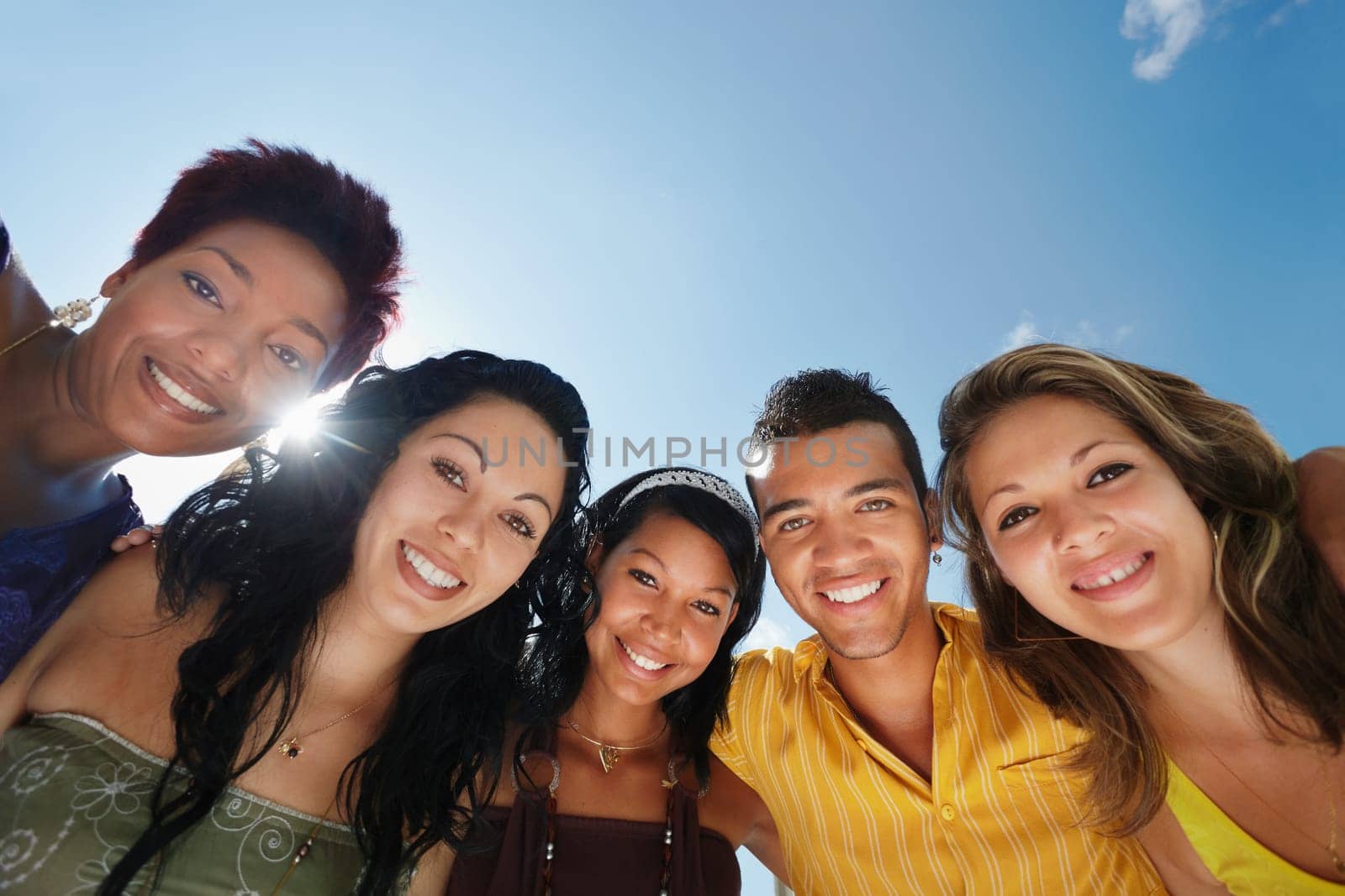 multiethnic group of five male and female friends hugging and looking at camera with sky in background. Low angle view, copy space