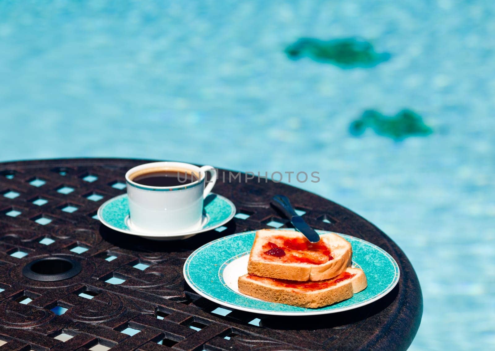 Coffee and plate on cast aluminum table and single chair by the side of swimming pool in back yard