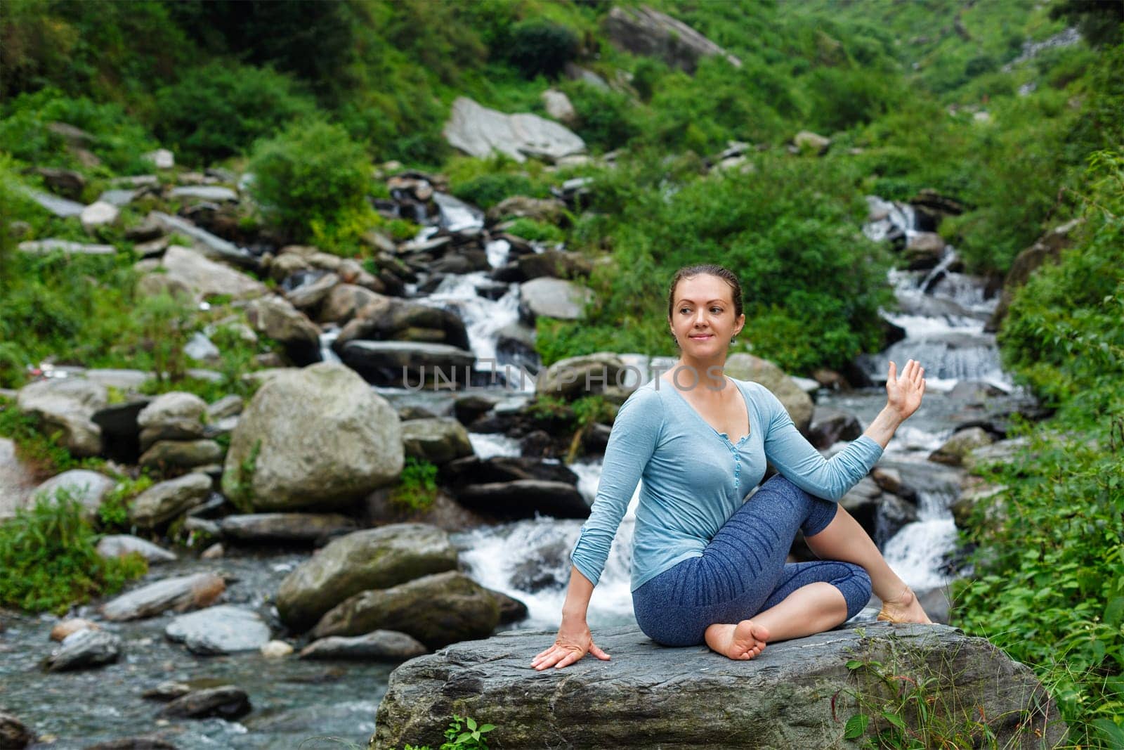 Yoga exercise outdoors - woman doing Ardha matsyendrasana asana - half spinal twist pose at tropical waterfall in Himalayas in India