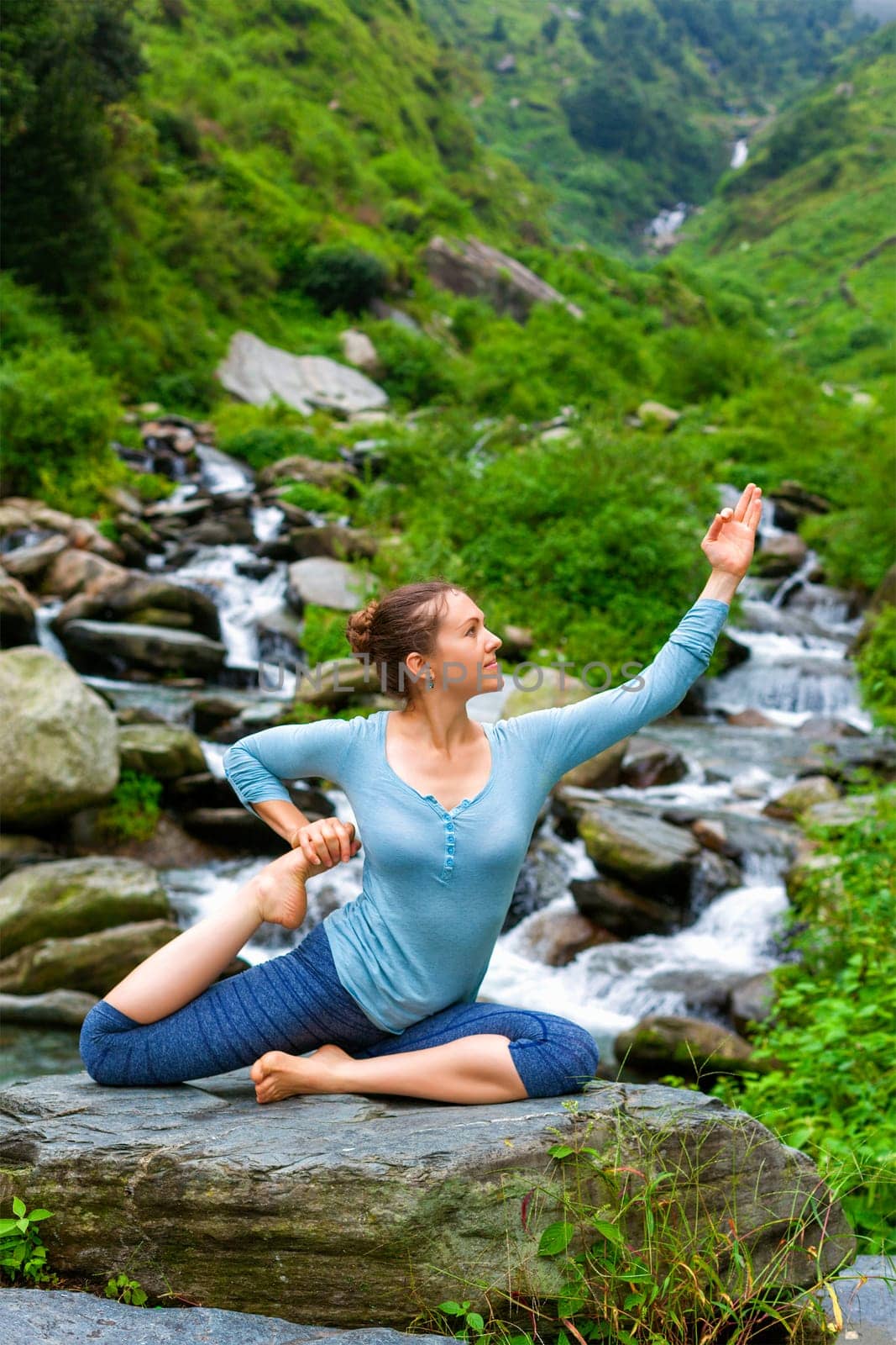 Hatha yoga outdoors - young sporty fit woman doing yoga asana Eka pada rajakapotasana - one-legged king pigeon pose at tropical waterfall. Himachal Pradesh, India