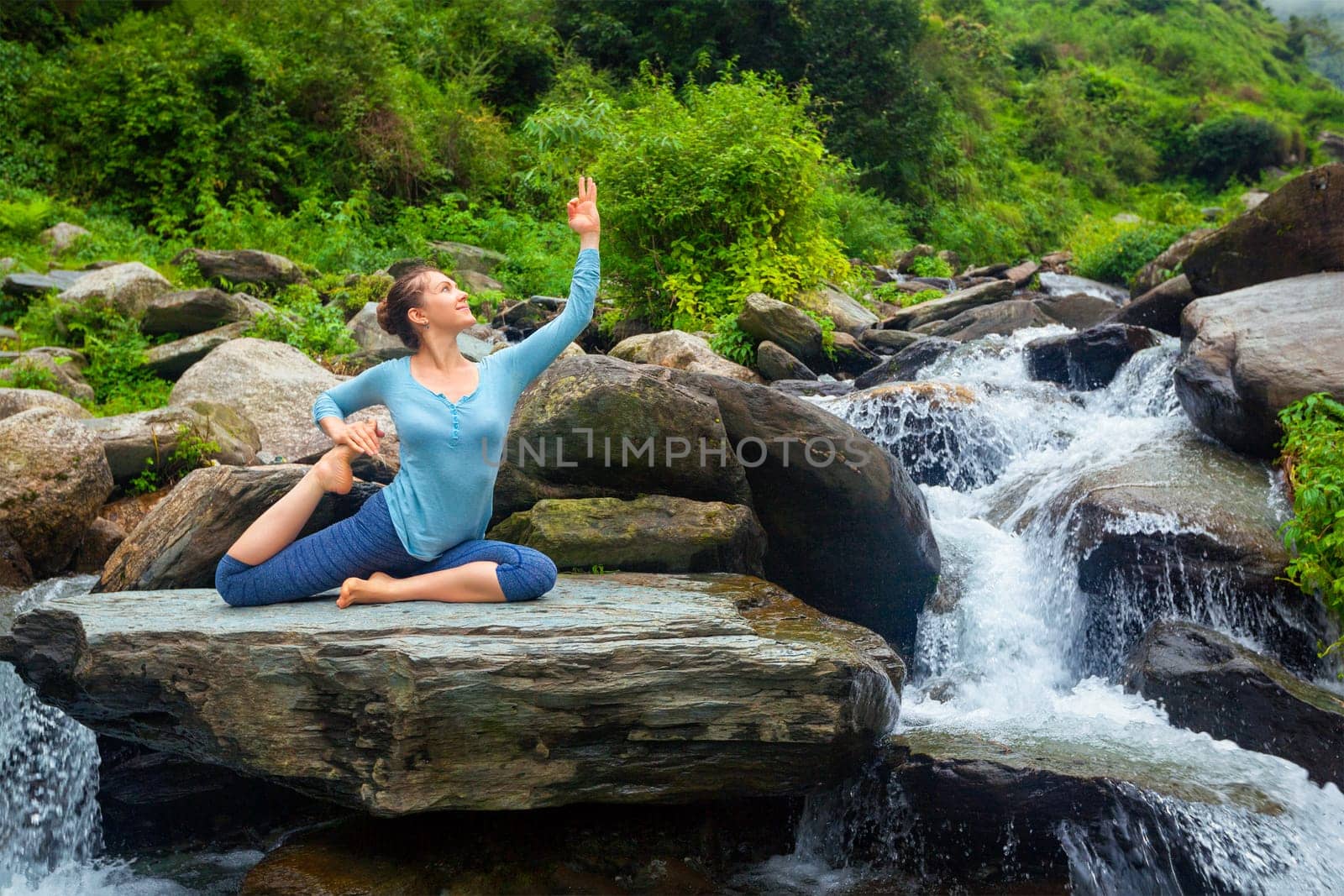Hatha yoga outdoors - young sporty fit woman doing yoga asana Eka pada rajakapotasana - one-legged king pigeon pose at tropical waterfall. Himachal Pradesh, India