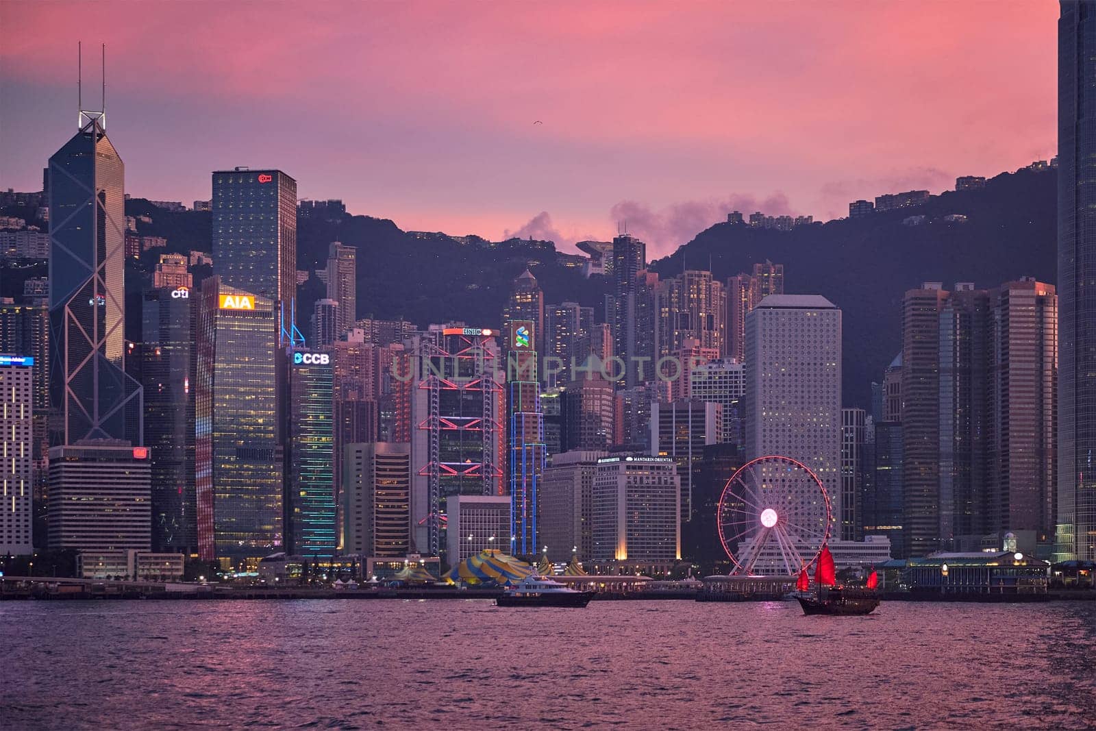 HONG KONG, CHINA - MAY 1, 2018: Tourist junk boat ferry with red sails and Hong Kong skyline cityscape downtown skyscrapers over Victoria Harbour in the evening. Hong Kong, China