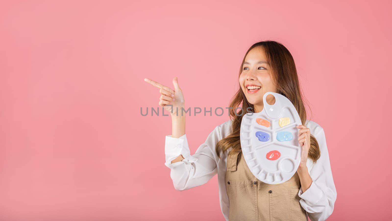 Portrait Asian young woman artist holding brush and paint palette, Happy female painting using paintbrush and palette with colors, studio shot isolated on pink background, Art design workshop