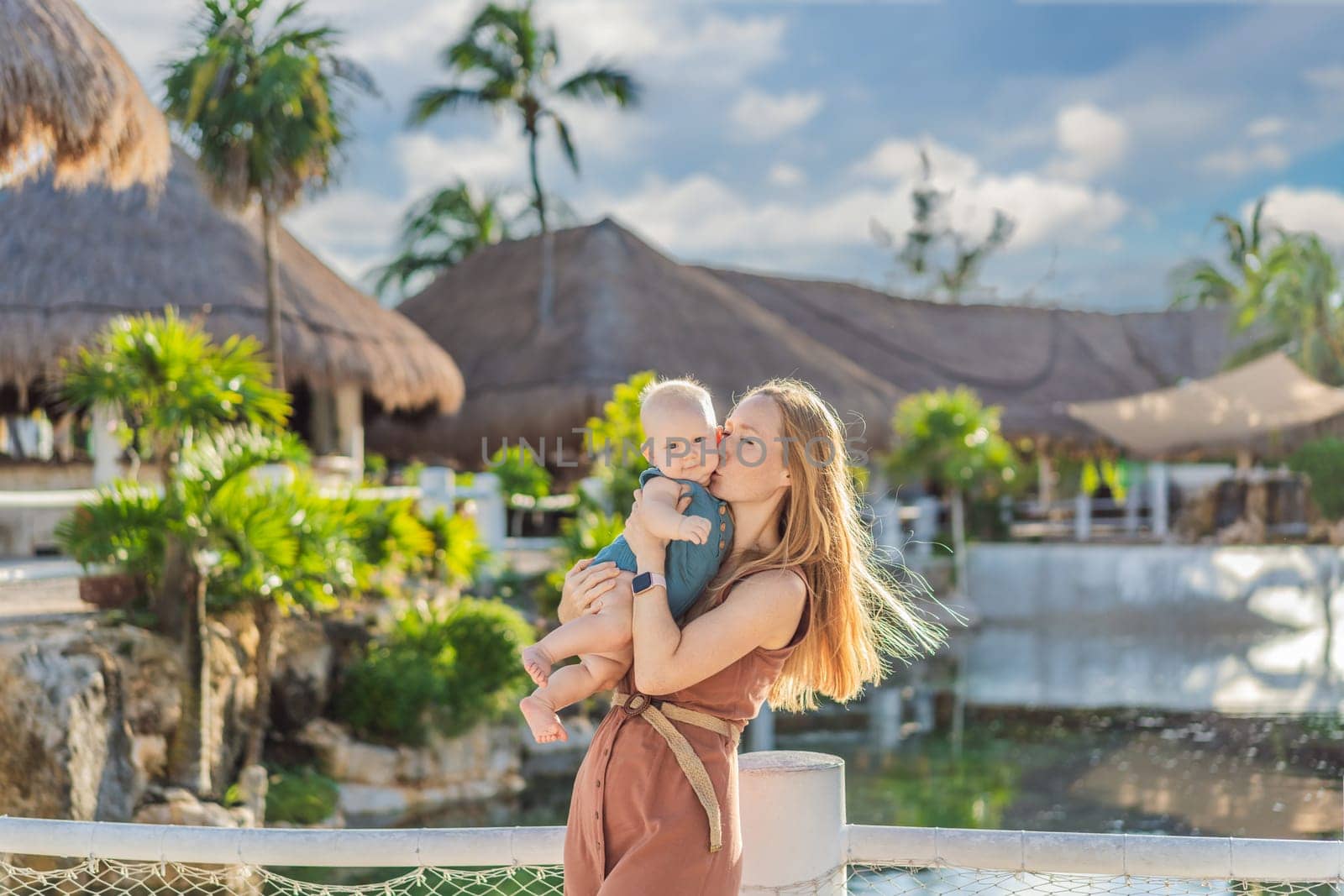 Amidst tropical palms and thatched roofs, a loving mom embraces her baby, sharing warmth and affection in a tranquil outdoor setting by galitskaya