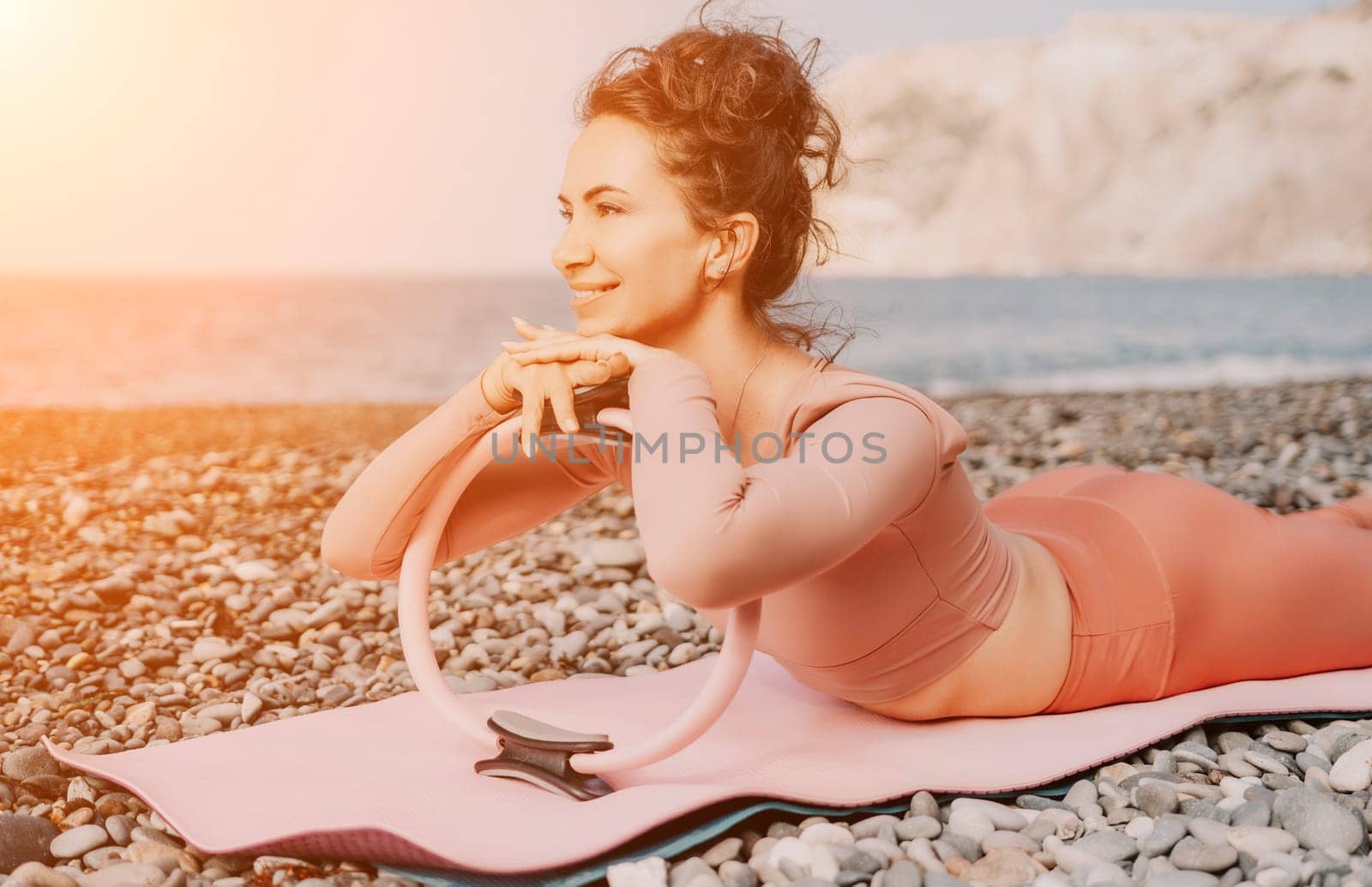 Middle aged well looking woman with black hair doing Pilates with the ring on the yoga mat near the sea on the pebble beach. Female fitness yoga concept. Healthy lifestyle, harmony and meditation.