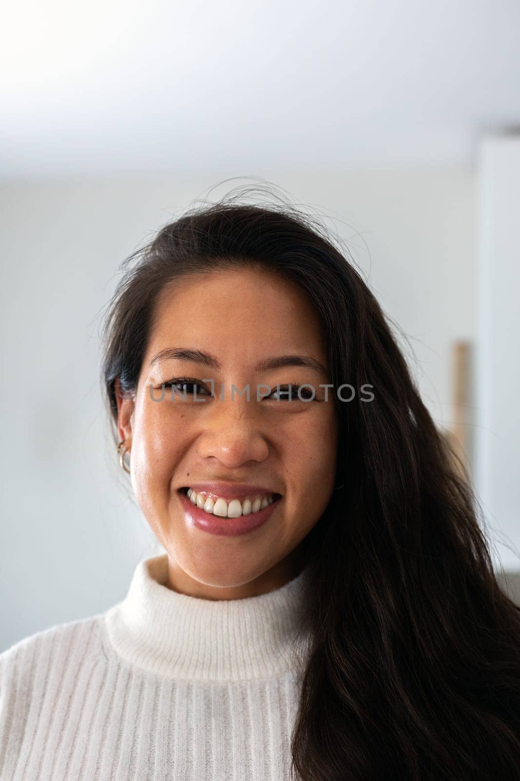 Vertical headshot of young smiling asian woman looking at camera indoors by Hoverstock