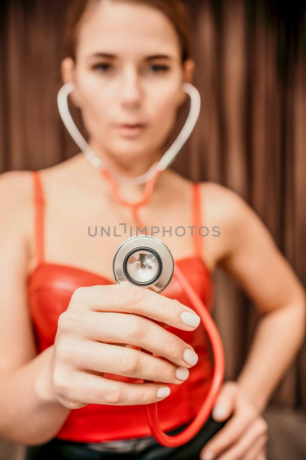Young woman in red corset and black leather pants with a phonendoscope. Sexy brunette nurse in red and black costume with a stethoscope in her hands. Role doctor play. Entertainment