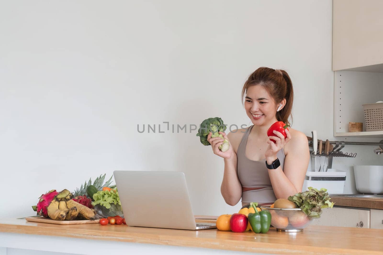 Beautiful Asian woman in workout clothes searches for healthy recipes online on her laptop. while preparing healthy food in the kitchen by wichayada