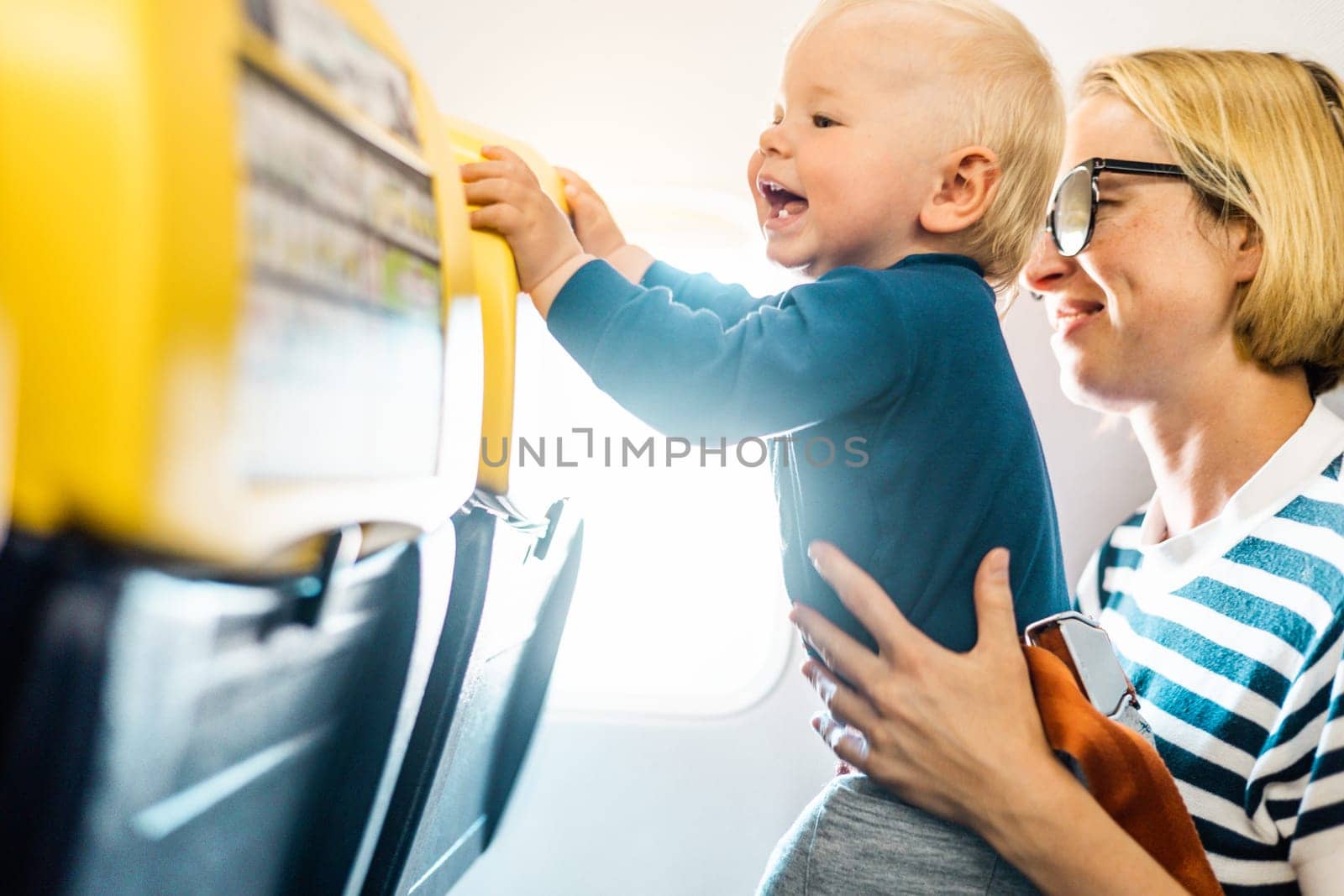 Mom and child flying by plane. Mother holding and playing with her infant baby boy child in her lap during economy comercial flight. Concept photo of air travel with baby. Real people
