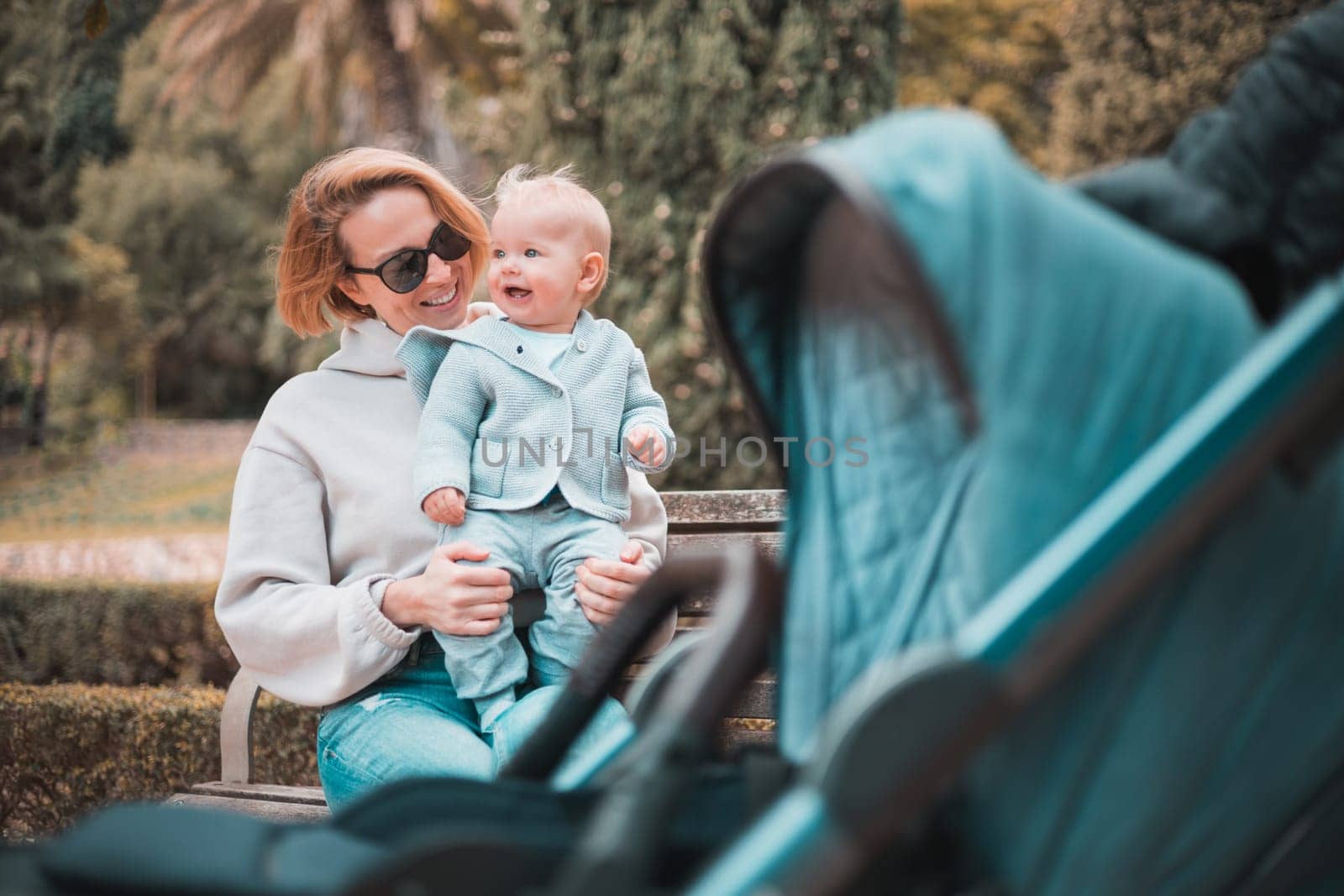 Mother sitting on bench in urban park, laughing cheerfully, holding her smiling infant baby boy child in her lap having baby stroller parked by their site. by kasto