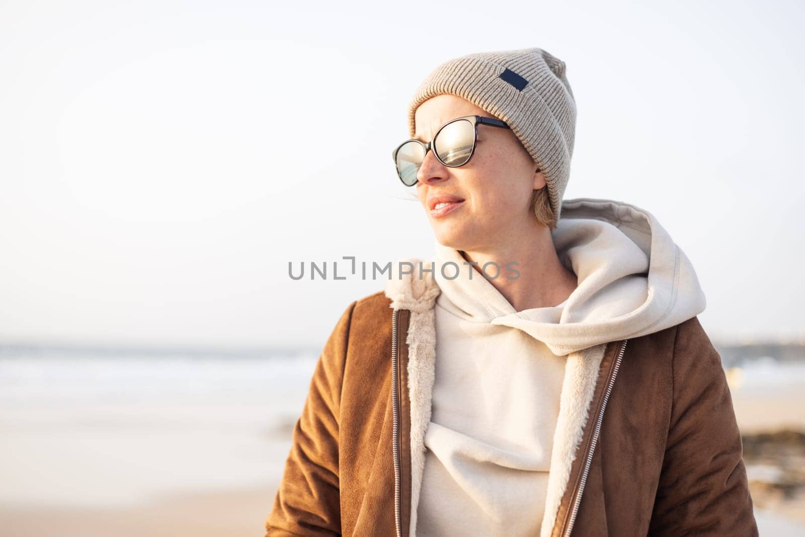 Portrait of young stylish woman wearing brown padded jacket, hoodie, wool cap and sunglasses on long sandy beach in spring. by kasto
