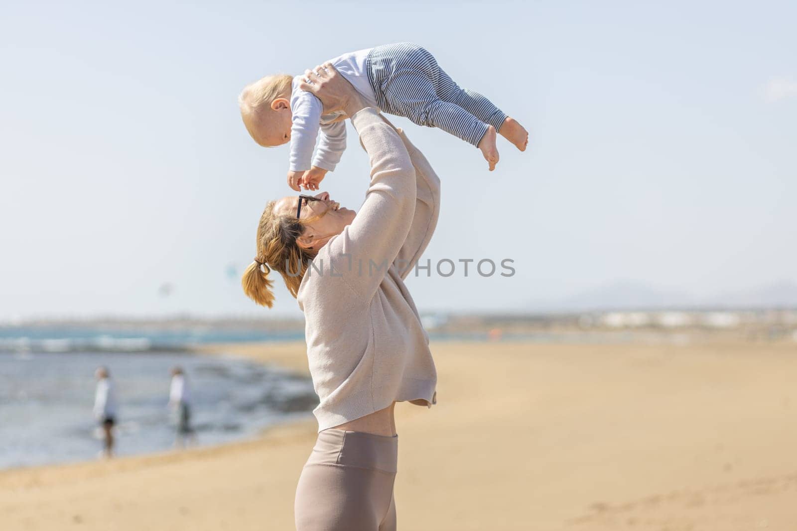 Mother enjoying summer vacations holding, playing and lifting his infant baby boy son high in the air on sandy beach on Lanzarote island, Spain. Family travel and vacations concept. by kasto