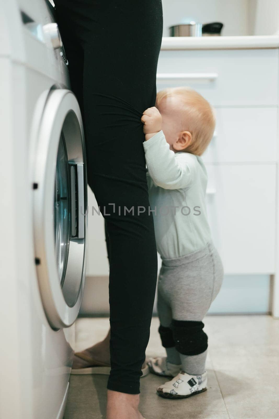 Little infant baby boy child hiding between mothers legs demanding her attention while she is multitasking, trying to do some household chores in kitchen at home. Mother on maternity leave