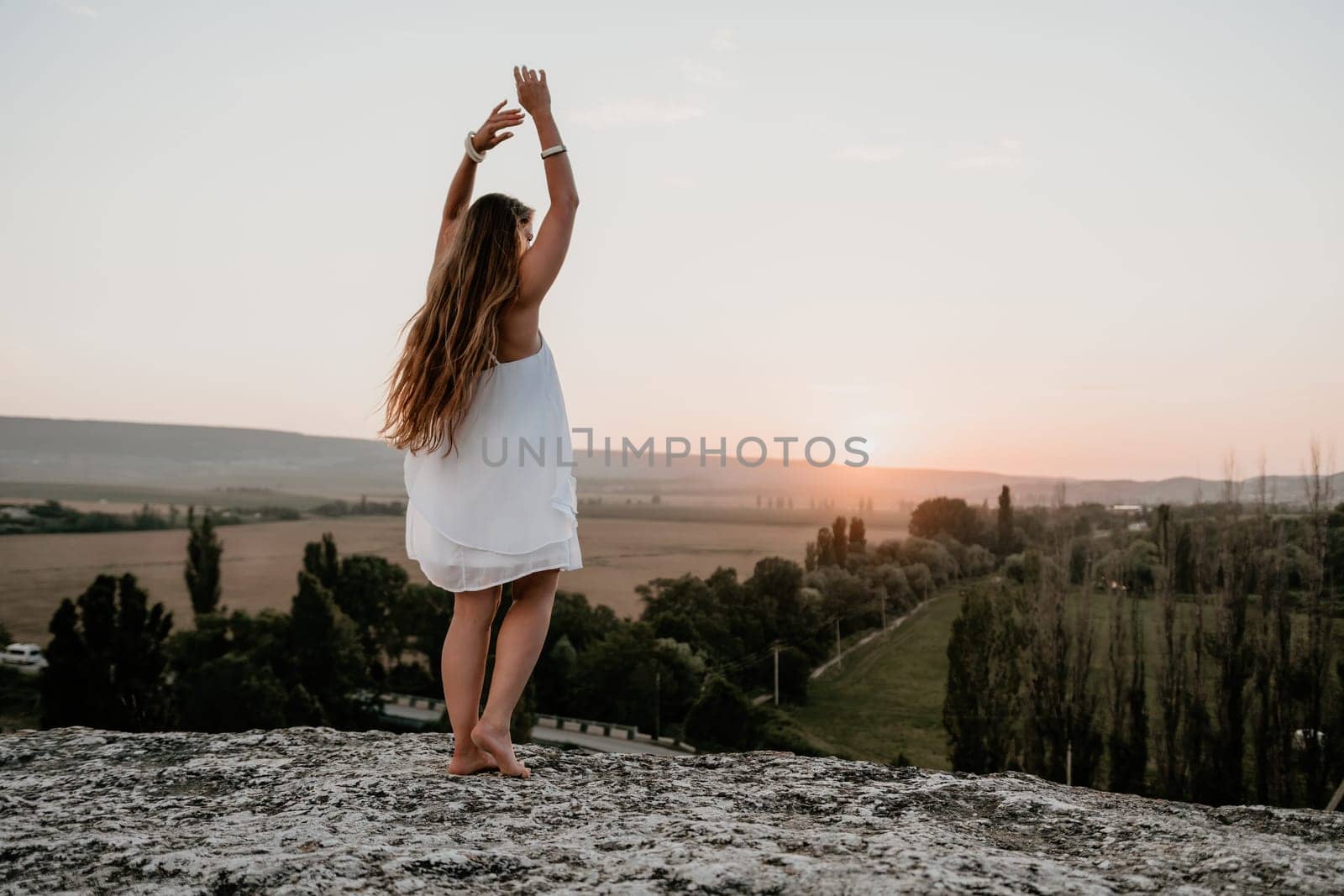 Happy woman standing with her back on the sunset in nature in summer with open hands. Romantic beautiful bride in white boho dress posing with mountains on sunset by panophotograph