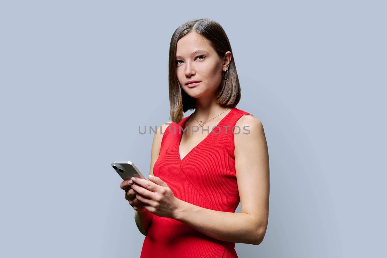 Young fashionable modern woman holding smartphone in hands, looking at camera on gray studio background. Technology, mobile applications, work leisure education lifestyle communication, youth people