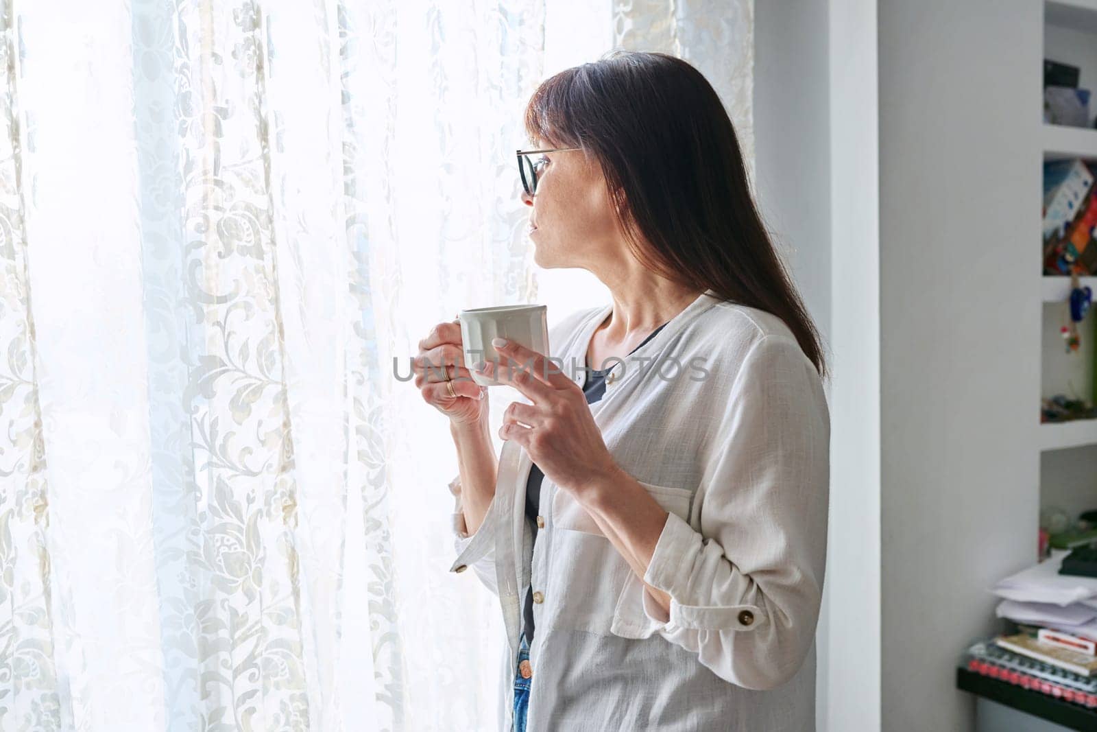 Serious middle-aged woman at home looking out window, with cup in her hands. Calmness, relaxation, loneliness, mature age people concept