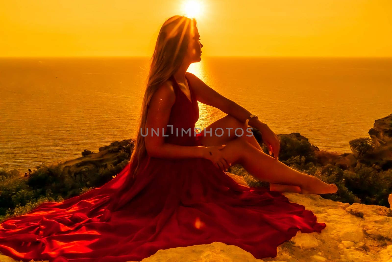 Woman sunset sea red dress, side view a happy beautiful sensual woman in a red long dress posing on a rock high above the sea on sunset. by Matiunina