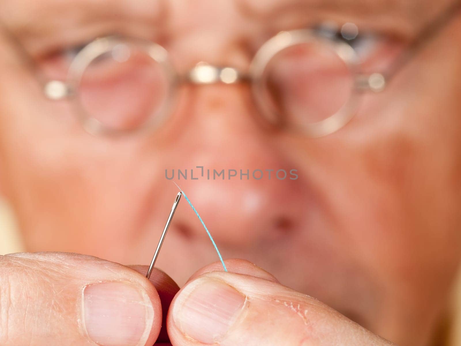 Macro image of a needle and cotton thread as senior man with glasses tries to thread the cotton
