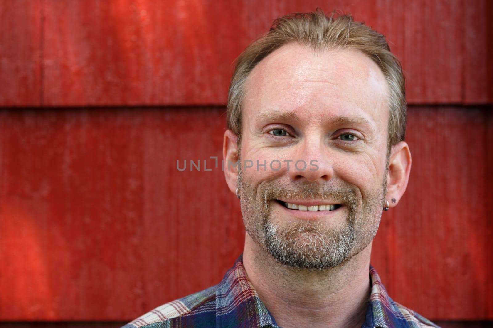 Happy man looking into the camera with a red house behind him in the day.