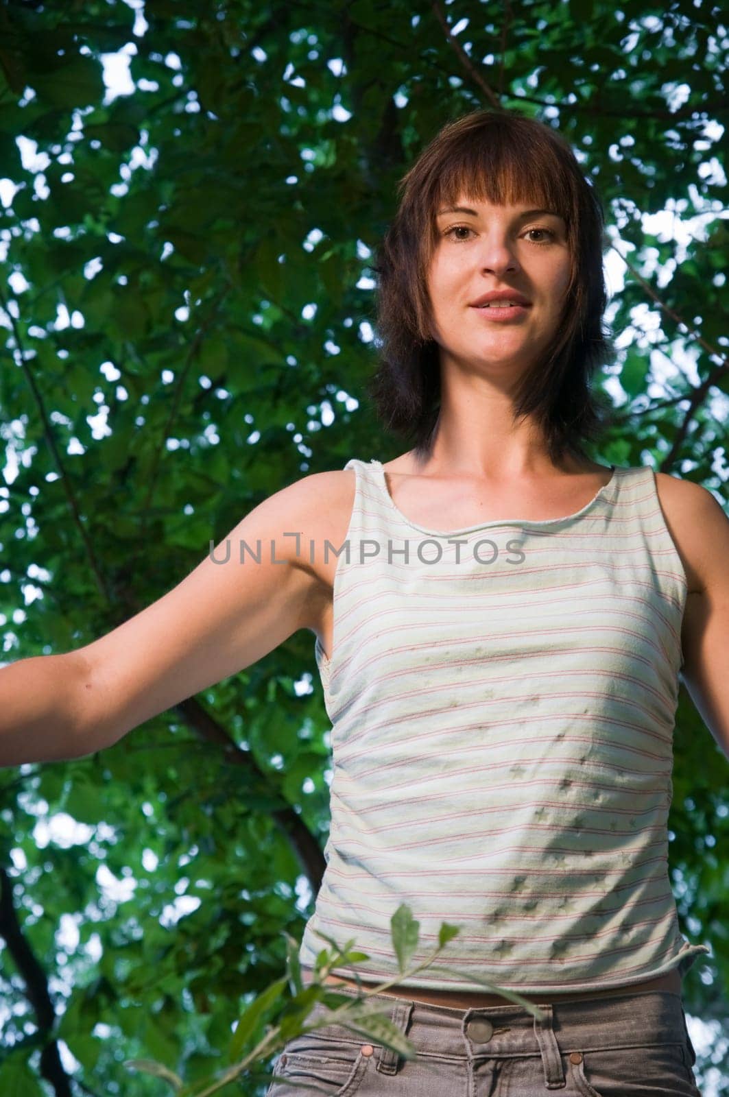 Upper body shot of a young brunette woman with medium length hair and sleeveless top and green tree leaves in the background