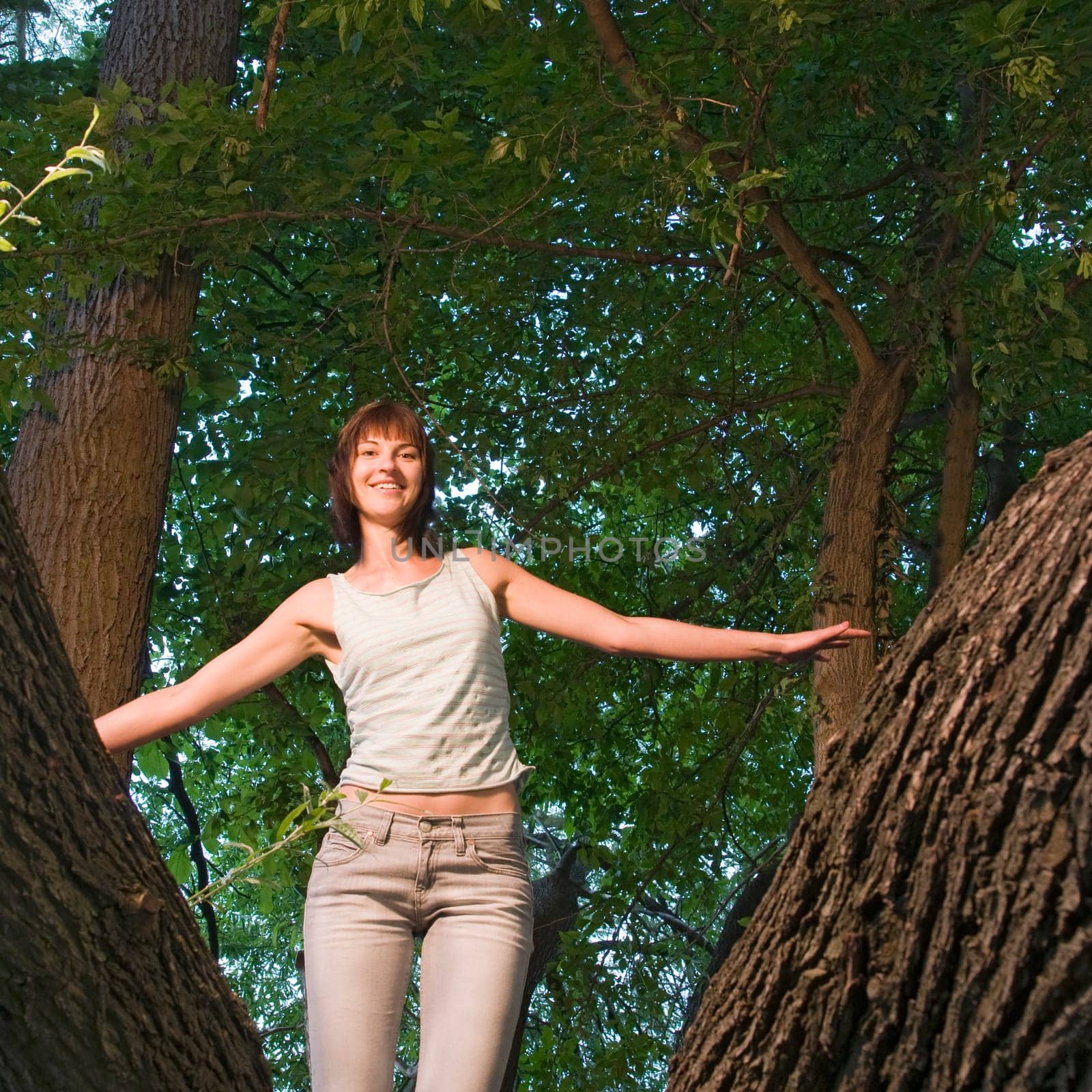 Three-quarter shot of a young brunette woman with medium-length hair and sleeveless top,arms outstretched standing in a forked branch with green tree leaves as a background