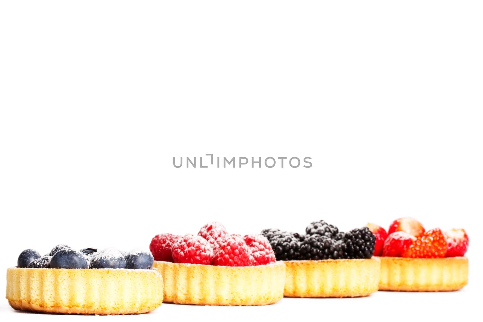 Sugared blueberries on tartlet cake in front of other berries on white background