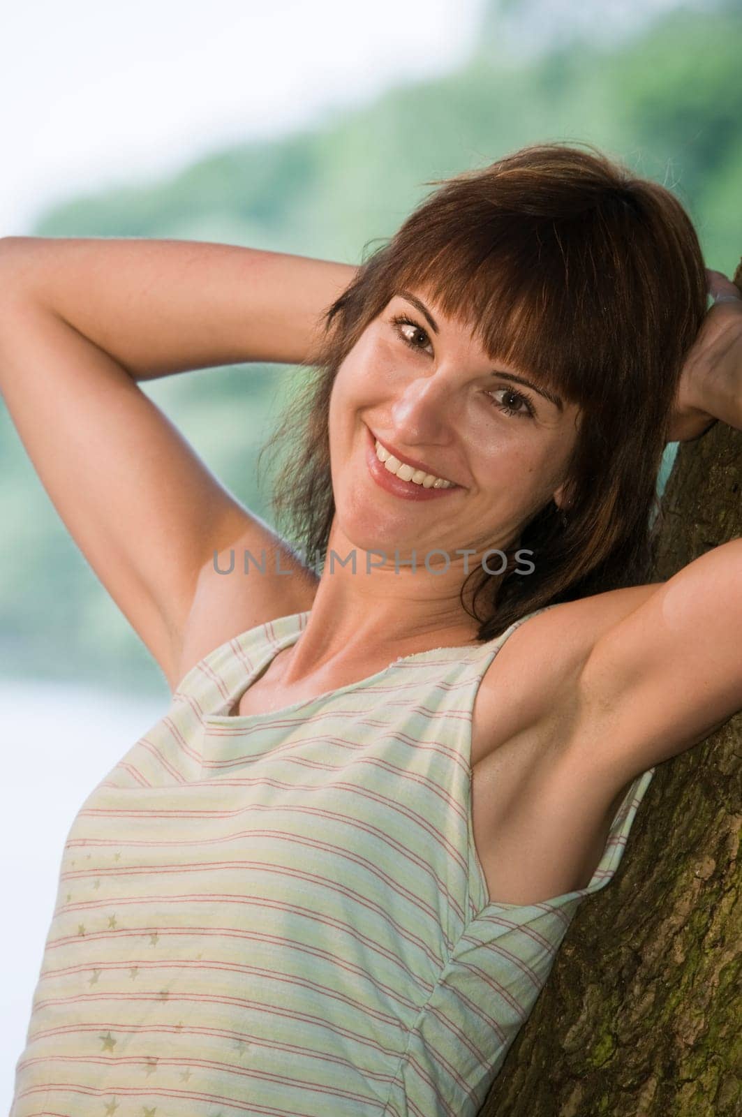 Upper body shot of a young woman leaning against a tree at a lake (Griebnitzsee in Potsdam / Germany) at sunset looking at the camera
