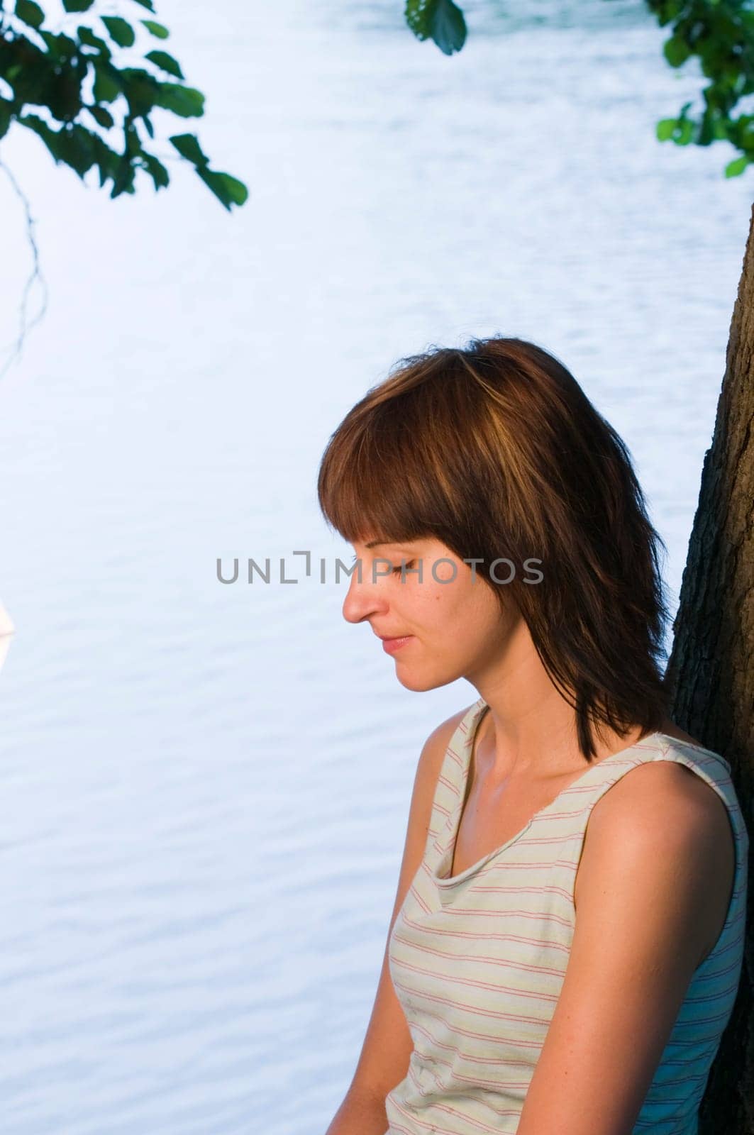 Side torso shot of a young woman in a short top by a lake and lowered thoughtful gaze,leaning against a tree trunk