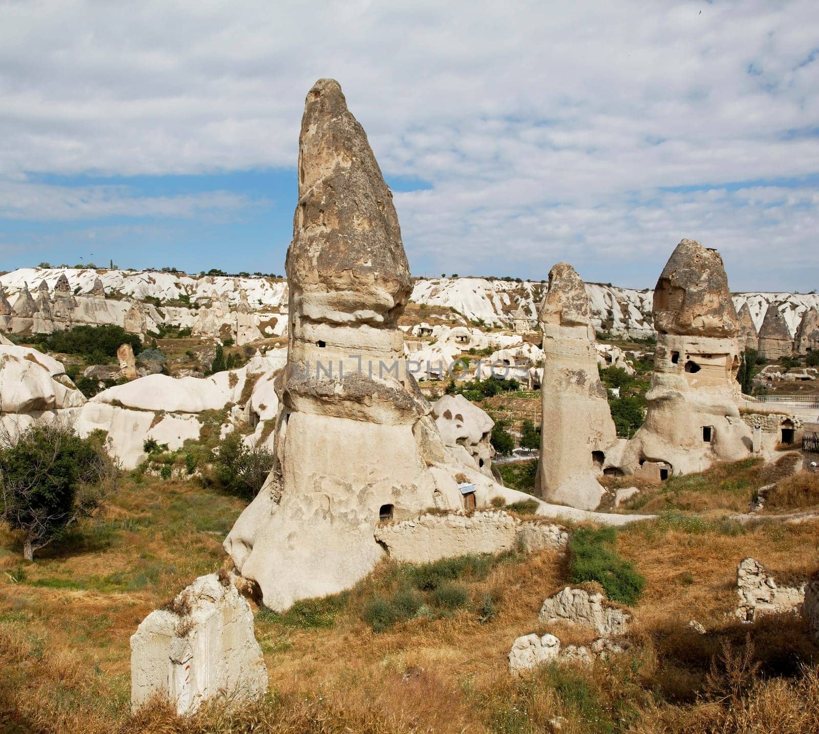 square format group of limestone fairy chimney homes near Goreme Cappadocia Turkey, volcanic lava hills in the background, cloudy blue sky with crop space and copy space