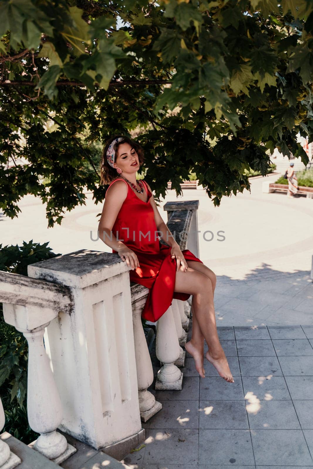 woman in a red silk dress and a bandage on her head smiles against the background of the leaves of a tree. She is leaning on the coop and looking into the camera. Vertical photo. by Matiunina