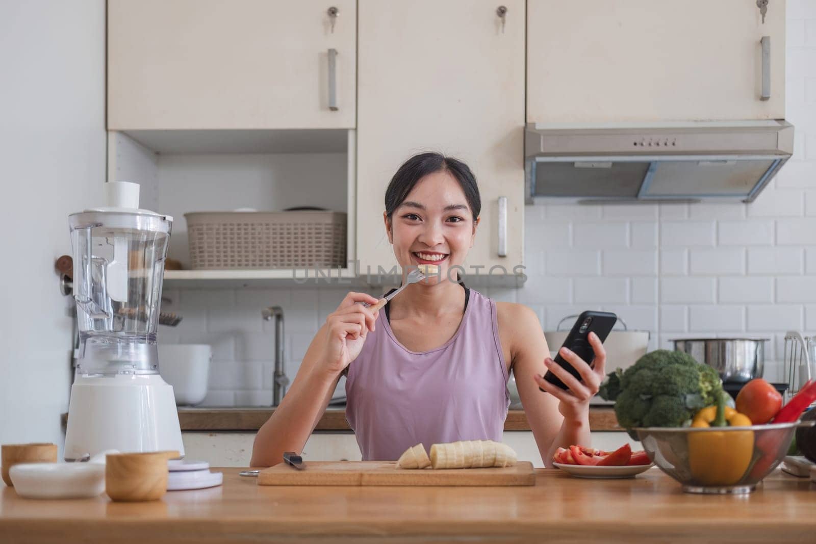 woman with casual clothes making salad and using mobile to count calories and plan diet. Happy woman consulting recipe instructions on smartphone while chopping vegetables in modern kitchen.