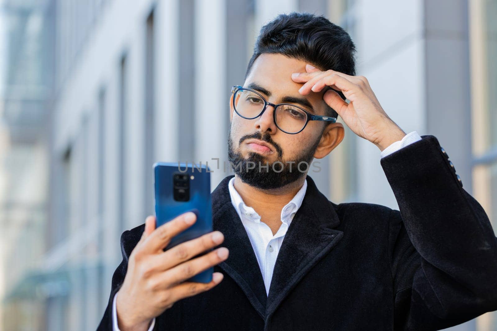 Indian businessman use smartphone typing browsing, loses surprised by bad news, fortune loss, fail bankruptcy outdoors. Arabian Hindu freelancer guy in downtown city street. Business people lifestyle