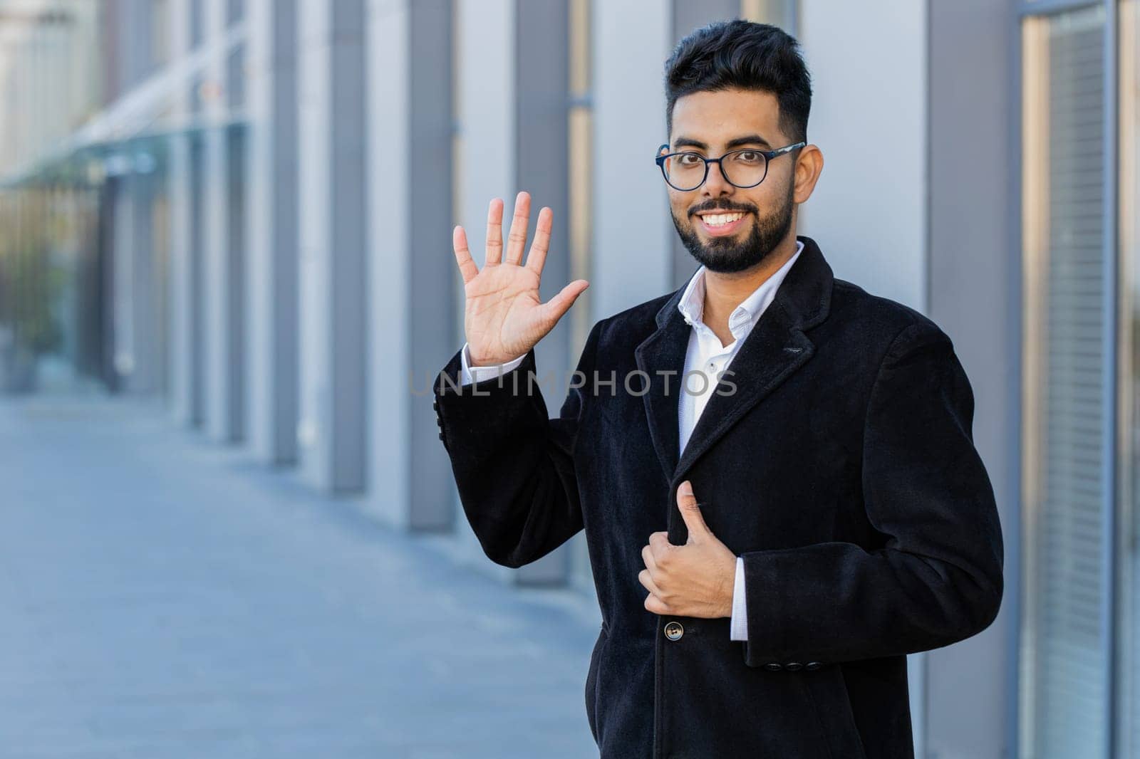 Hello. Indian businessman smiling friendly at camera, waving hands gesturing hi, greeting or goodbye welcoming with hospitable expression outdoors. Arabian Hindu freelancer guy in downtown city street