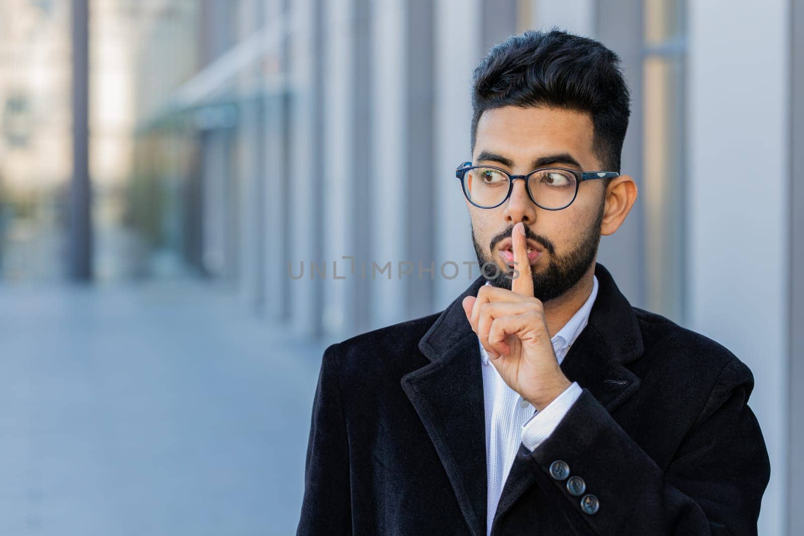 Shh be quiet please. Indian businessman presses index finger to lips makes silence hush gesture sign do not tells gossip secret outdoors. Arabian Hindu guy in downtown city. Business people lifestyles