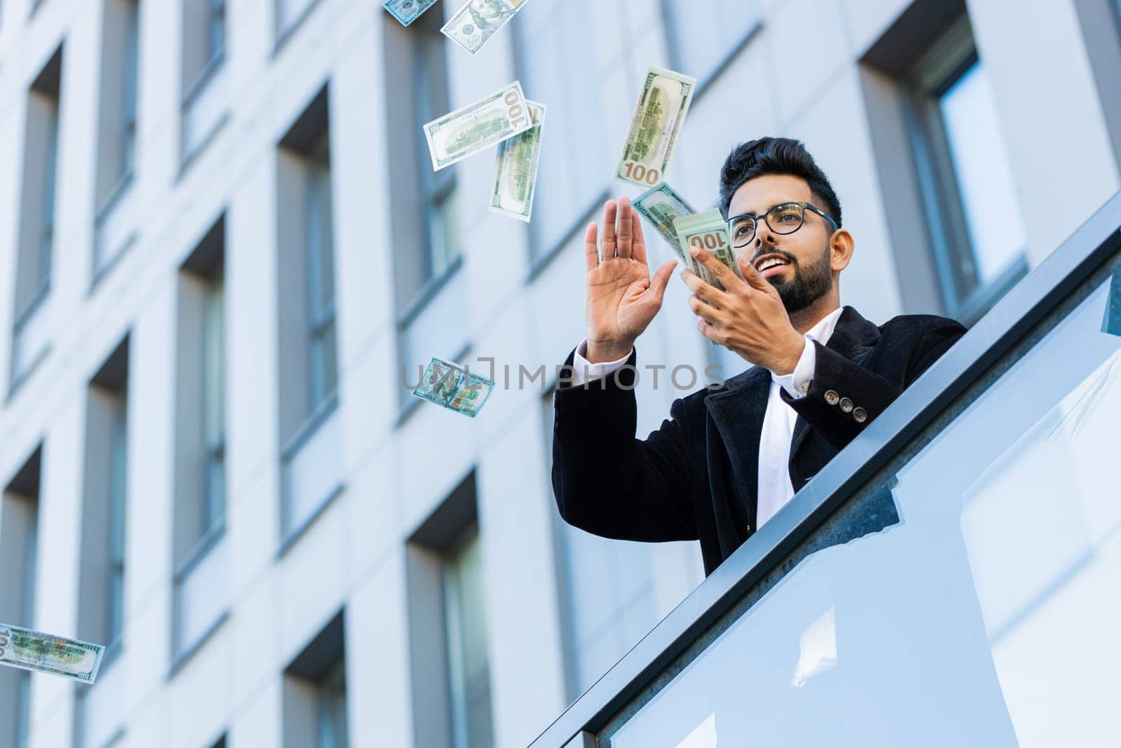 Happy rich Indian businessman wasting throwing money dollars cash around earnings, big profit, win lottery, share, celebrate, charity donation outdoors. Arabian Hindu freelancer guy in downtown city