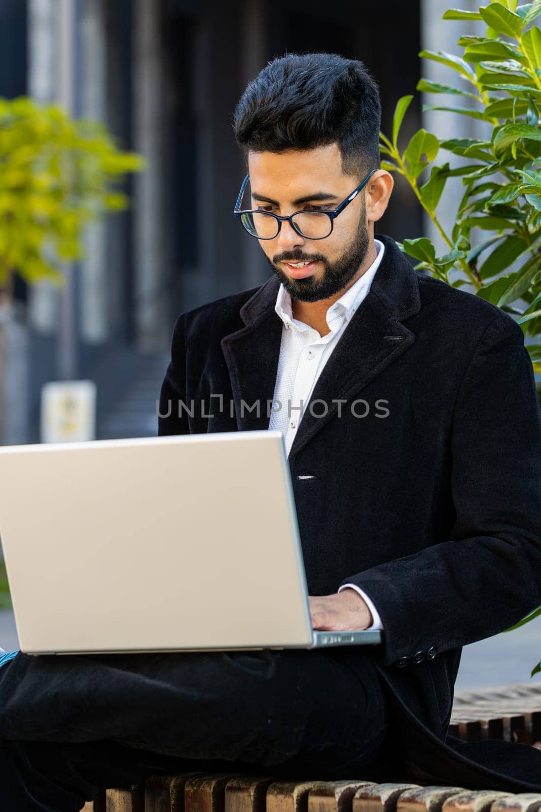 Happy Indian businessman typing working remote on wireless laptop computer browsing online chat app outdoors. Arabian Hindu freelancer office guy in downtown city street. Business people. Vertical