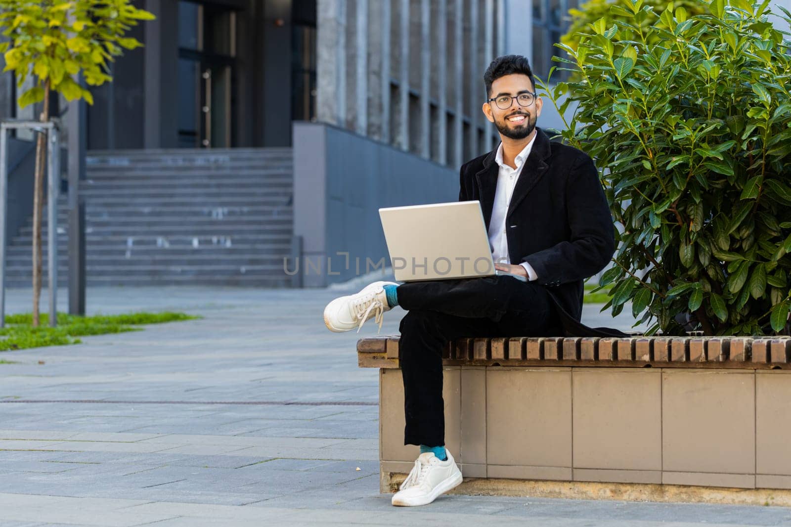 Happy Indian businessman typing working remote on wireless laptop computer browsing online chat app outdoors. Arabian Hindu freelancer office guy in downtown city street. Business people lifestyles
