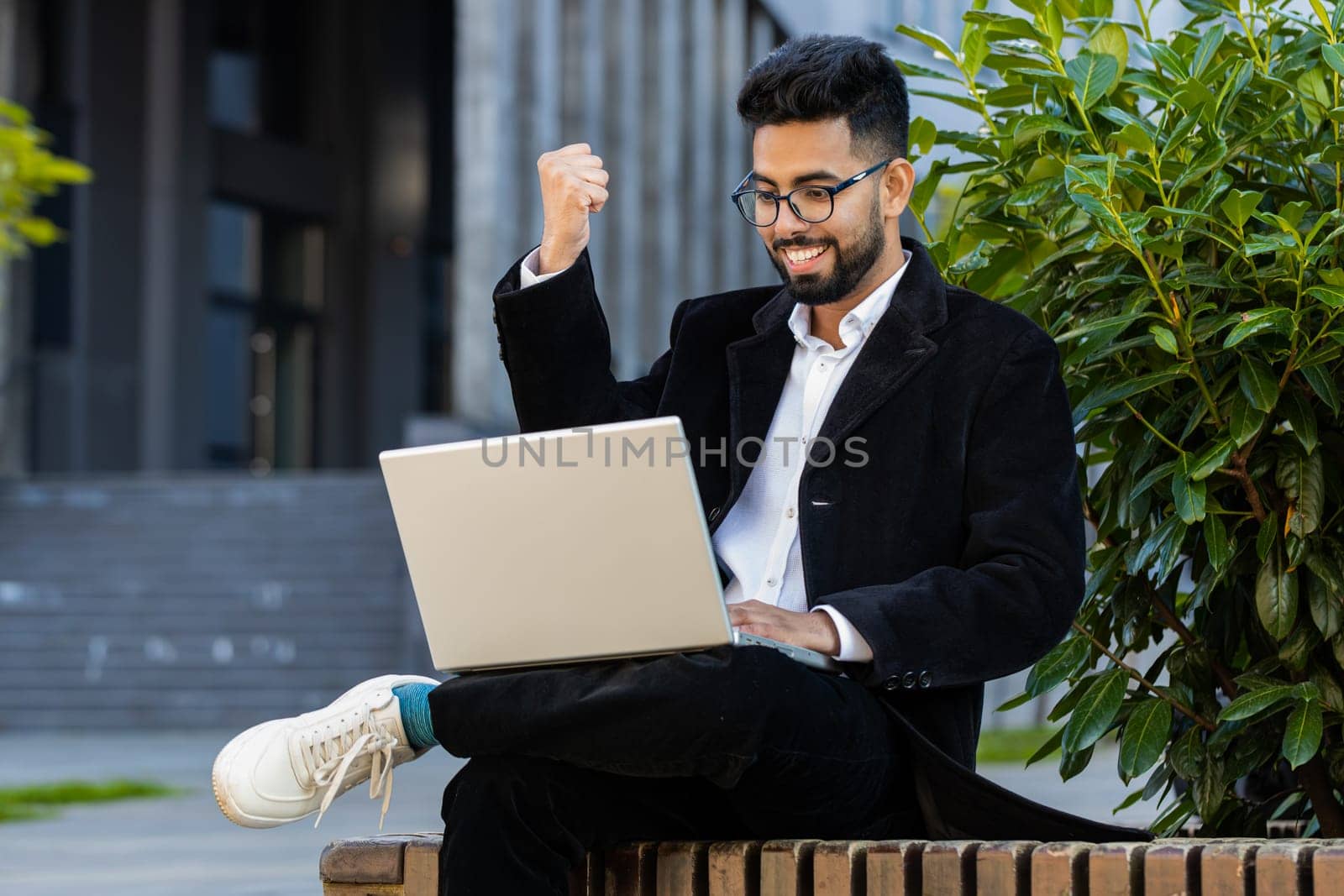 Indian businessman working on laptop reading great good news, rejoicing success, celebrate online win victory internet achievement prize outdoors. Arabian Hindu freelancer guy sitting in downtown city