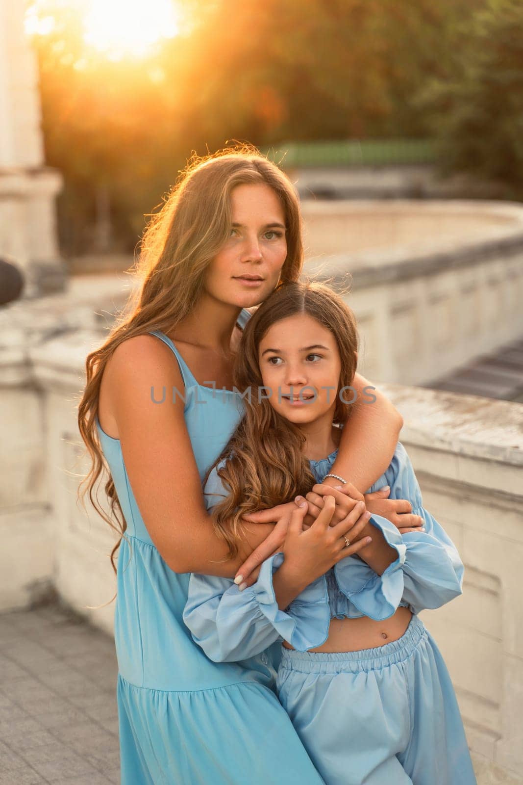 Portrait of mother and daughter in blue dresses with flowing long hair against the backdrop of sunset. The woman hugs and presses the girl to her. They are looking at the camera