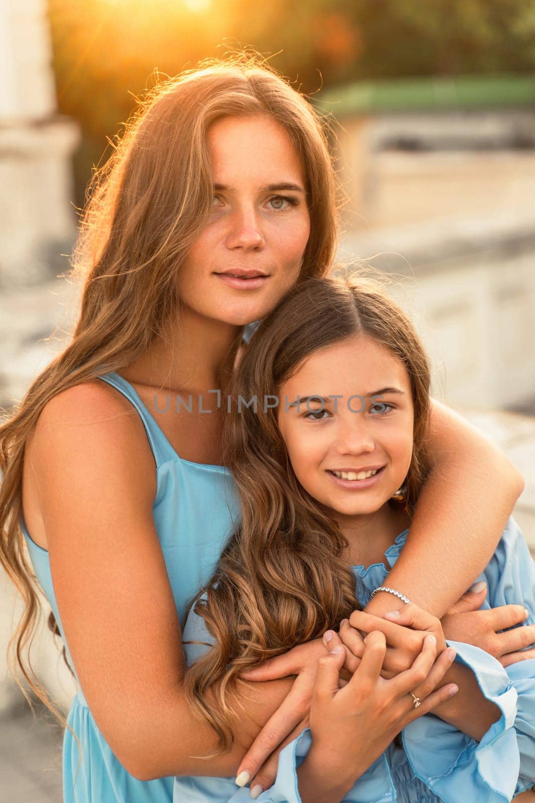 Portrait of mother and daughter in blue dresses with flowing long hair against the backdrop of sunset. The woman hugs and presses the girl to her. They are looking at the camera. by Matiunina