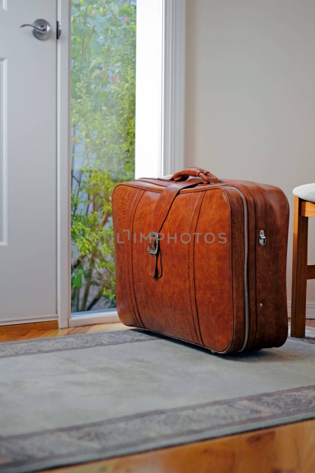 One brown suitcase sitting on the entrance rug inside the front door of a home in the daytime waiting to be taken on some travel.