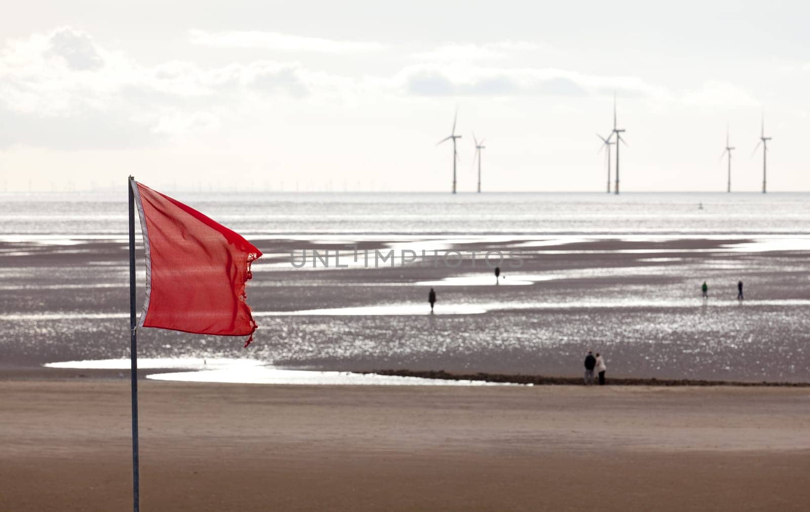 Red flag warns swimmers as wind turbines turn in the ocean off a sandy beach near Liverpool on a cold and cloudy day