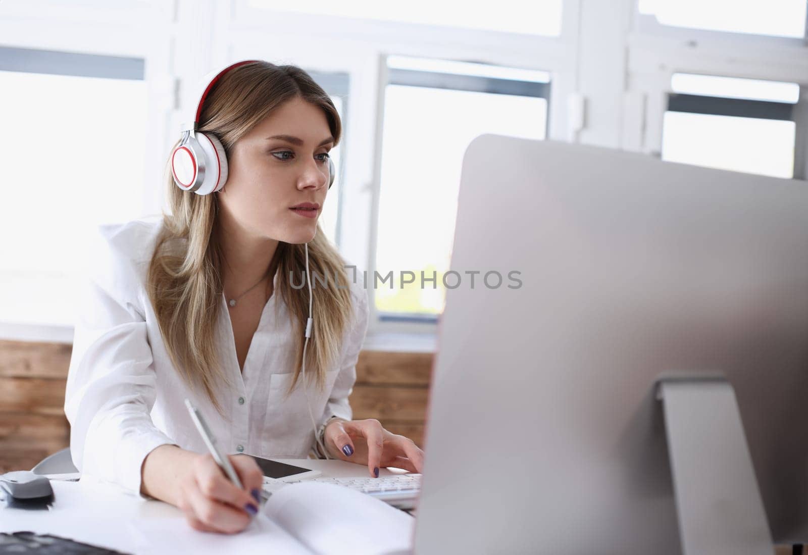 Beautiful woman wearing headphones making notes with silver pen while working on computer pc portrait. Management training course modern remote school self development and perfection concept