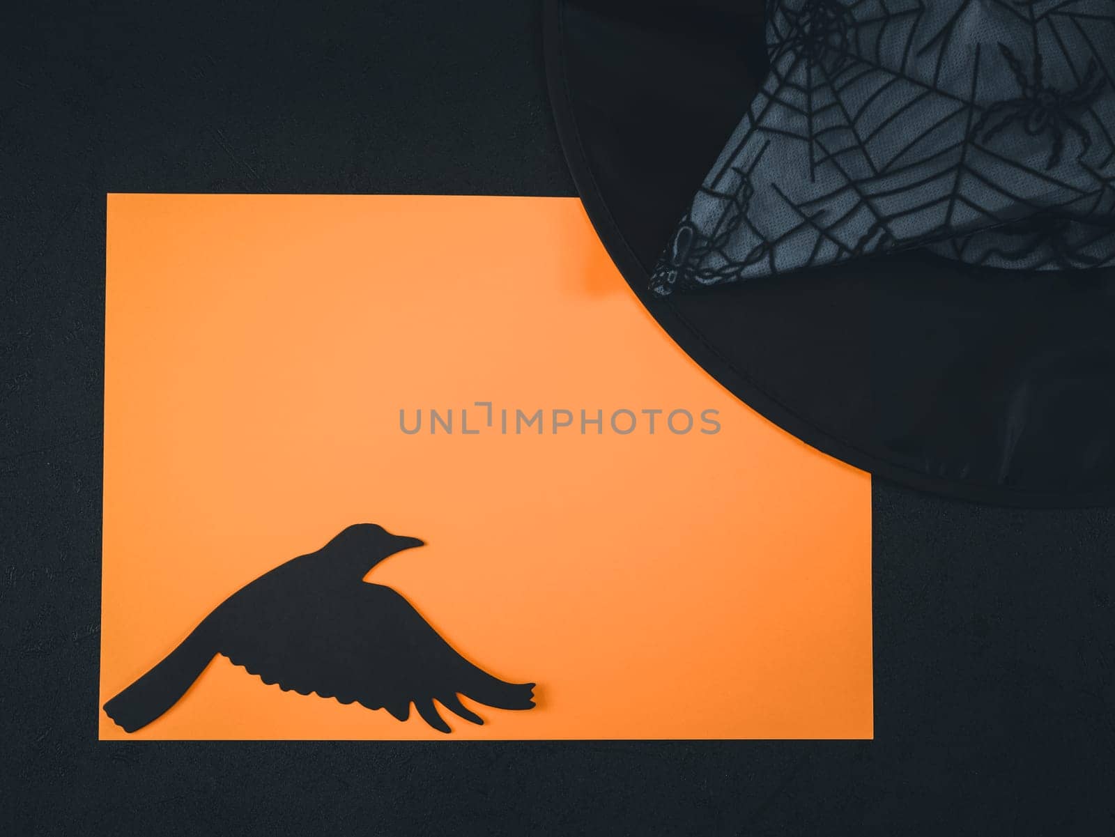 One blank orange sheet lies horizontally against a black background with a paper crow and a witch hat on the sides, flat lay close-up.