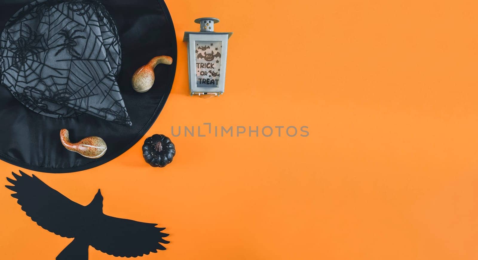 Witch hat, lantern, black paper crow and three decorative pumpkins lie on the left on an orange background with space for text on the right, flat lay close up. Scary concept, banner.