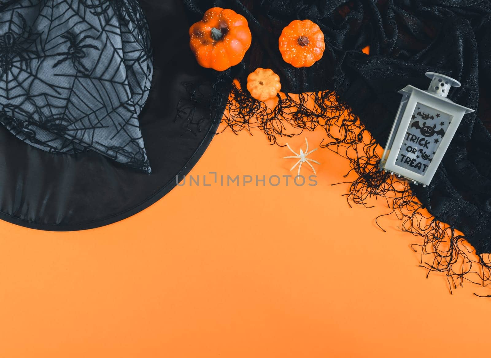 A witch hat, a lantern, a black shawl, a spider and three decorative pumpkins lie on top on an orange background with space for text at the bottom, flat lay close up. Scary party concept.
