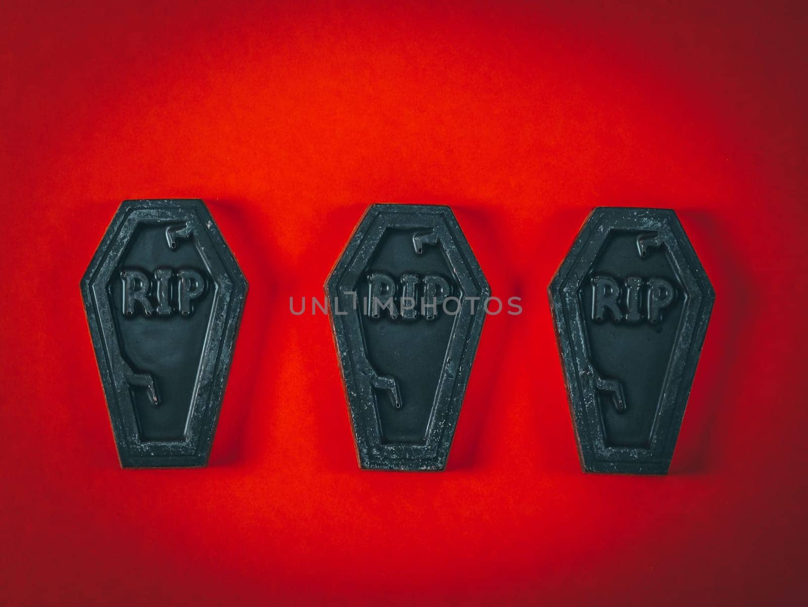 Three chocolate coffins with the words rip lying in a row in the center of a red-orange background with vignette around the edges, flat lay close-up. Scary halloween candy concept.